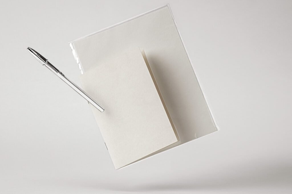 Blank white notebook, a clear plastic cover, and a silver pen floating against a plain light gray background, ready for branding.
