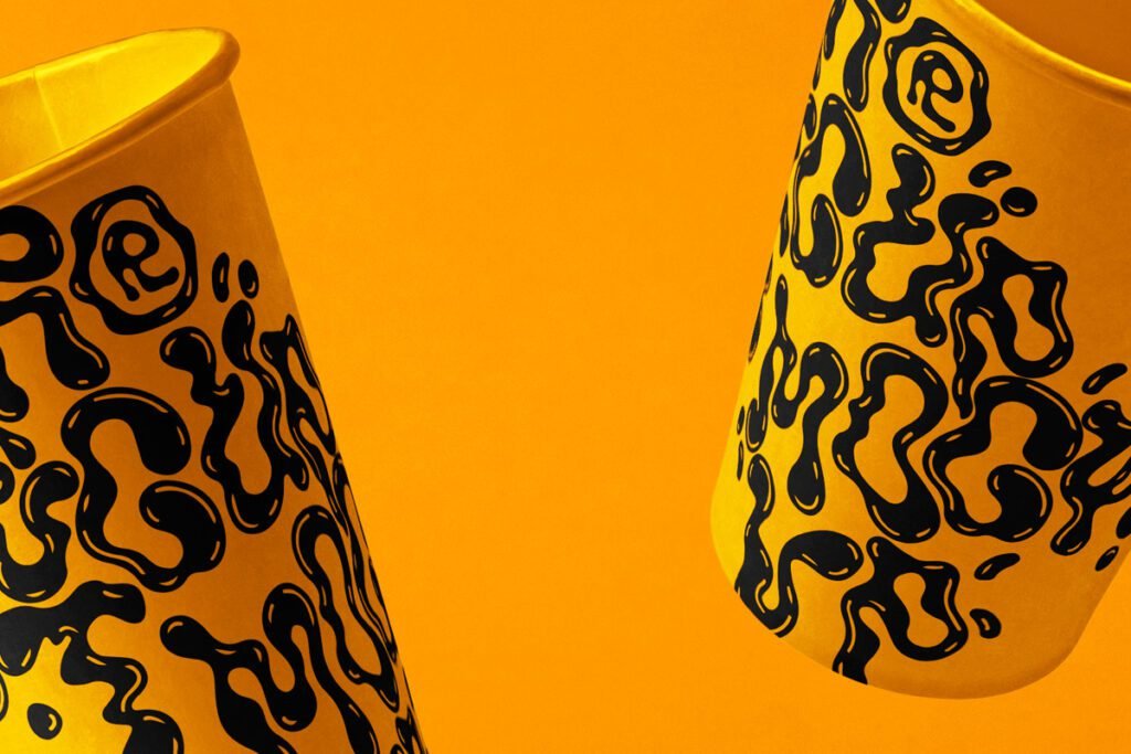 Two yellow disposable cups with black abstract liquid-drip patterns, angled and floating against a vibrant orange background, ideal for beverage branding.
