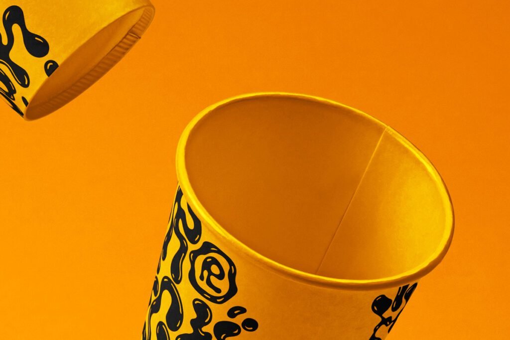Two yellow paper cups with black liquid-drip patterns appearing to float against a vibrant orange background, showcasing beverage branding.