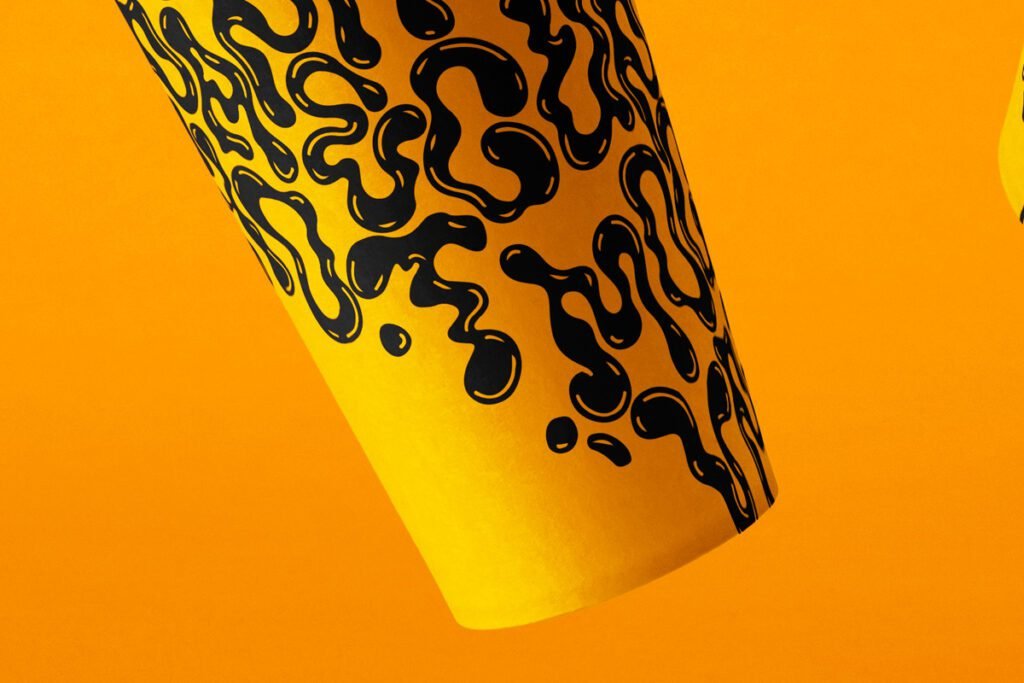 Close-up of a yellow paper coffee cup mockup with a bold black organic, liquid drip pattern, angled against a vibrant orange background.