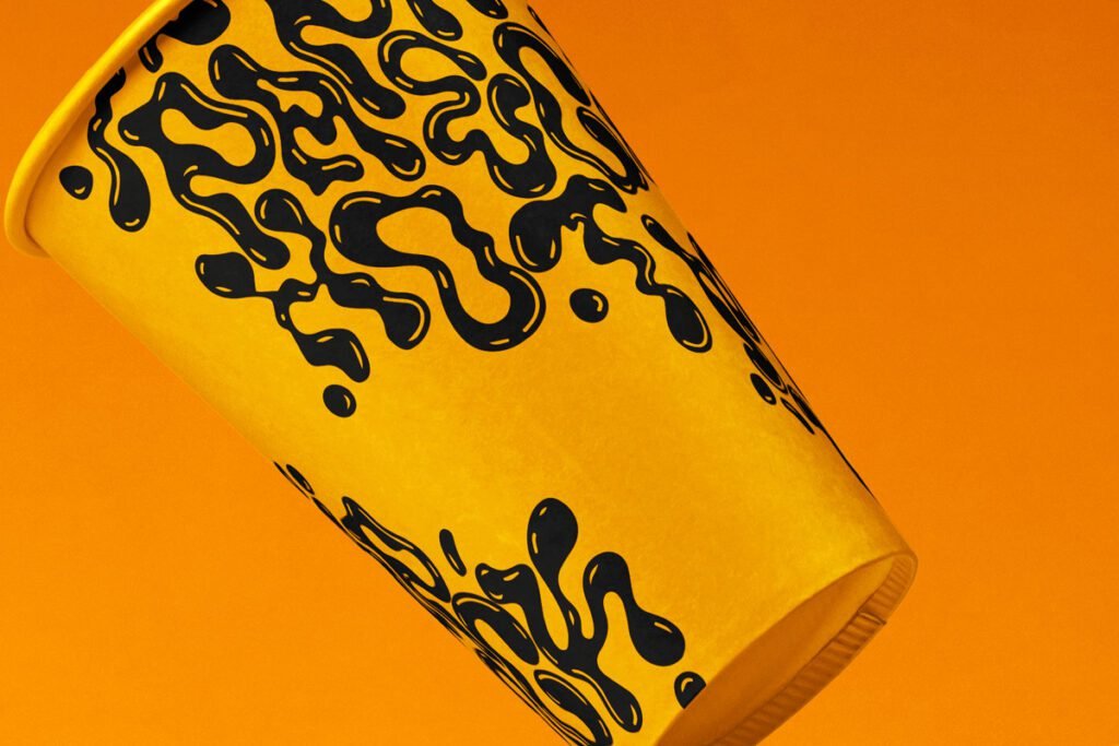 Vibrant yellow paper cup mockup with black liquid drip pattern, angled against a matching orange background, perfect for beverage branding.