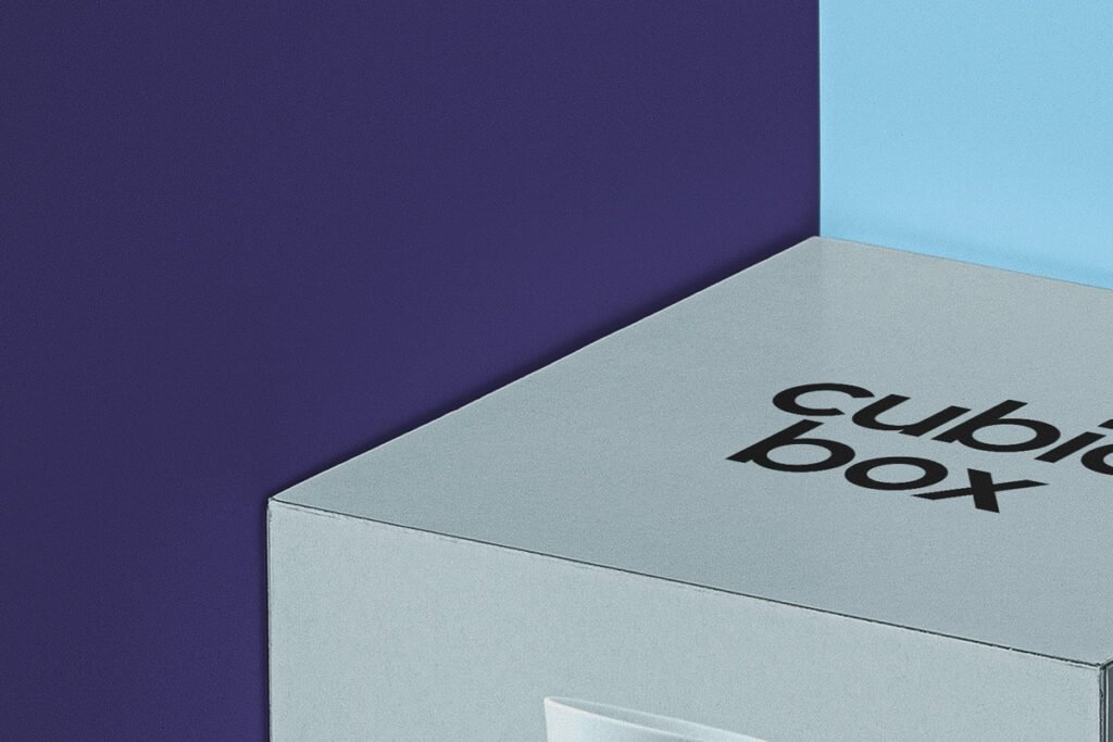 A close-up, high-angle view of the top and side of a light grey "Cubic Box Packaging" with "cubic box" branding in black, positioned against a corner backdrop of deep purple and light blue.
