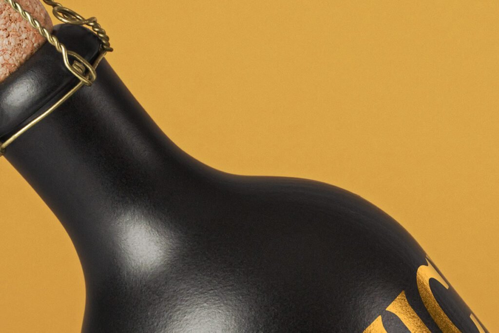 Close-up of a matte black bottle with a cork and gold wire cage, showing a hint of gold typography, set against a warm yellow background.