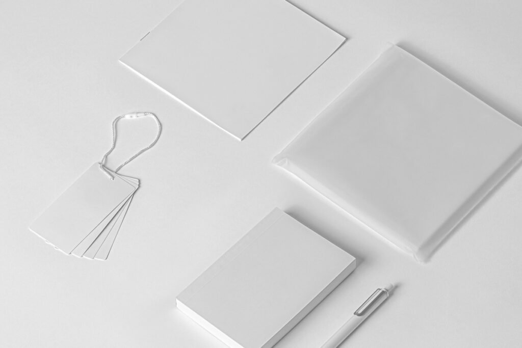 An overhead view of a clean, minimalist mockup set on a white background, featuring blank white hang tags with a string, two square flat items (possibly booklets or cards), a transparently wrapped item, and a white pen.