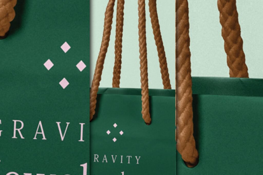 A close-up of a dark green paper bag mockup featuring a white geometric logo and part of the word "GRAVITY," with prominent brown rope handles.