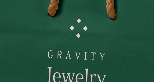 A close-up of a dark green paper bag mockup displaying "GRAVITY Jewelry" in elegant white and light pink fonts, topped with a geometric logo and visible brown rope handles.