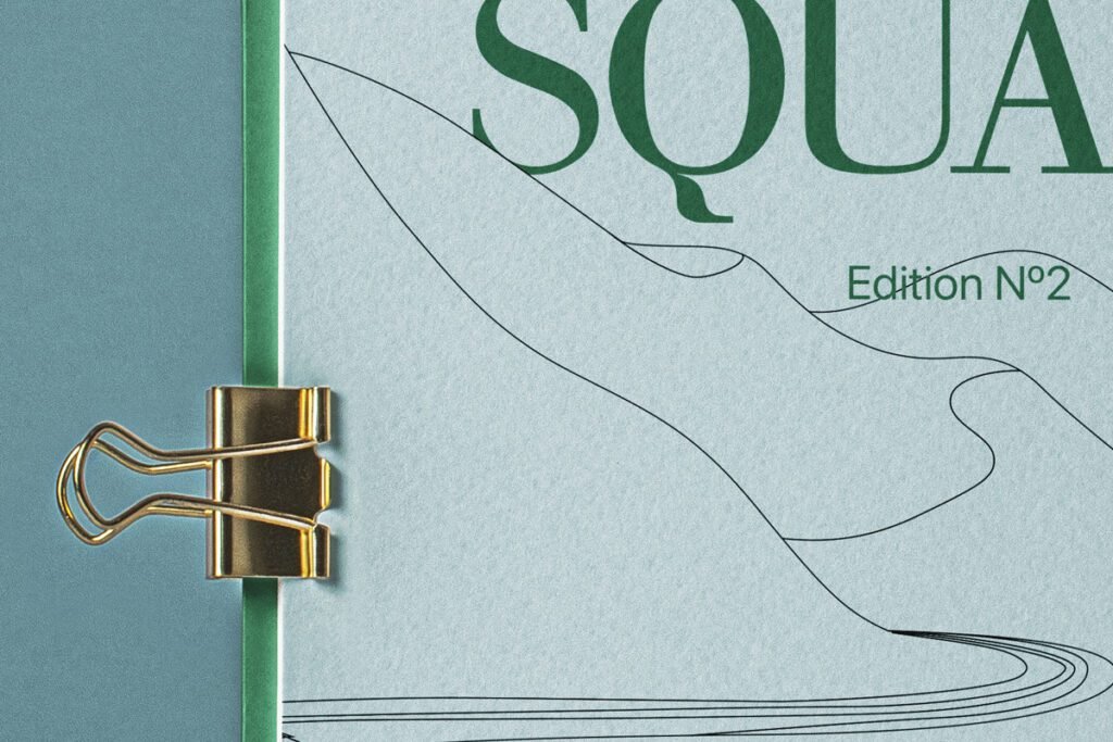 A close-up of a Square Catalog Mockup, featuring a light blue catalog with abstract black line art and "Edition N°2" in green text, bound by a golden binder clip to a dark teal cover.