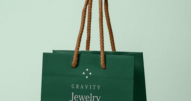 A dark green paper bag mockup held by its brown rope handles, featuring a "GRAVITY Jewelry" brand logo and text on a light green background.