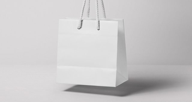 A blank white paper shopping bag with white rope handles, appearing to float slightly above a light white surface, against a plain white background.