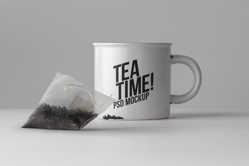 A white ceramic mug with black "TEA TIME! PSD MOCKUP" text is positioned on a light gray surface, next to a translucent pyramid-shaped tea bag filled with dark tea leaves.