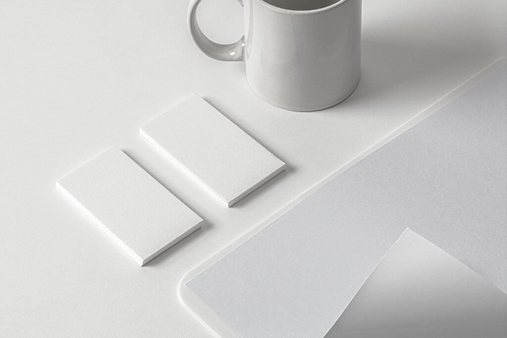 A minimalist Stationery Branding Mockup PSD featuring two blank white business card stacks, a light gray ceramic mug, and a stack of white papers on a clean white desk, ideal for clean branding presentations.