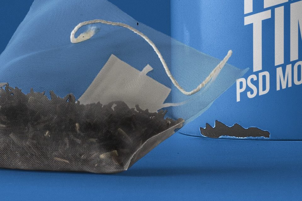 A close-up of a "Psd Tea Mug Mockup" on a blue background, showing a transparent tea bag filled with dark tea leaves in the foreground and the bottom right of a blue mug with white text partially visible.