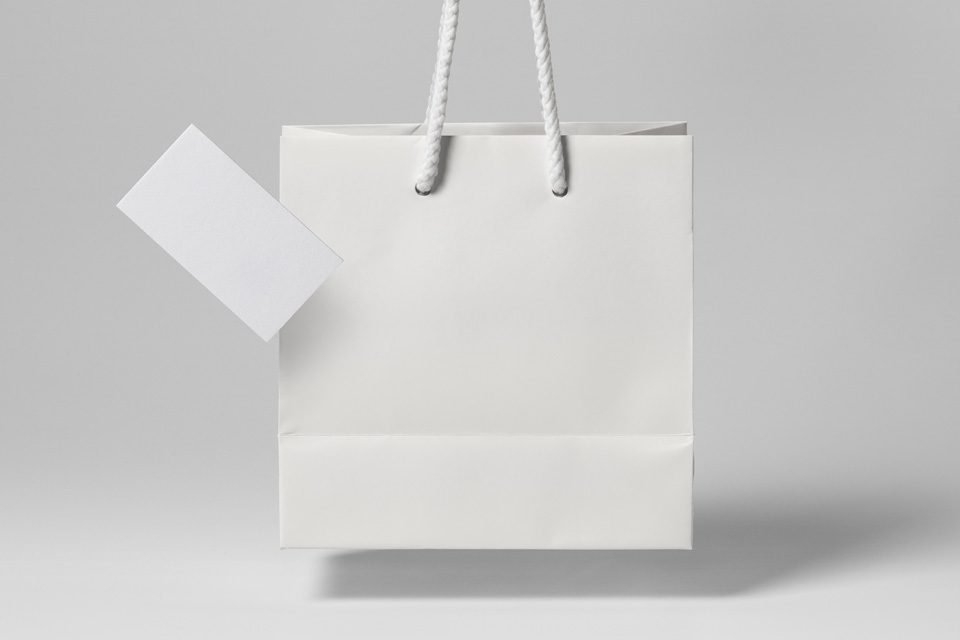 A floating white "Shopping Bag PSD" with white rope handles and a blank white rectangular tag protruding from its left, all against a plain light grey background.