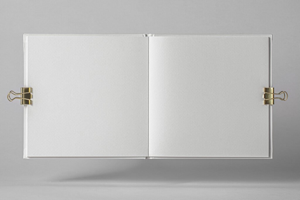 A top-down view of an open, blank square book or catalog mockup with white pages, held open by two gold binder clips on a light grey background.