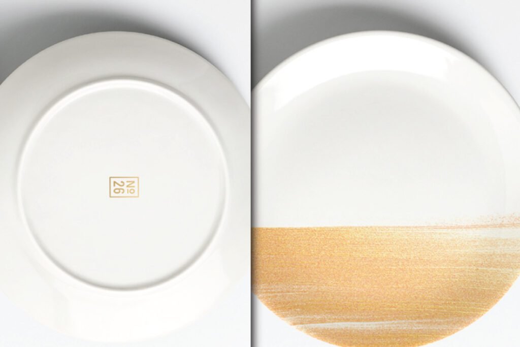 Best Realistic Ceramic Plate Wrap Mockup PSD showcases two white ceramic plates. One is seen from the back with a gold number 26, and the other from the front with a horizontal gold brush stroke design.