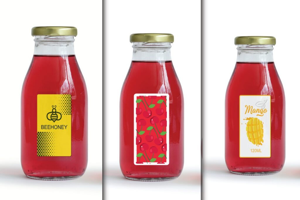 Juice and Honey Glass Bottle Label Mockup PSD showing three glass bottles with red liquid and gold caps. Labels include a yellow honey label with a bee, a white label with red cherries, and a white label with a mango design.