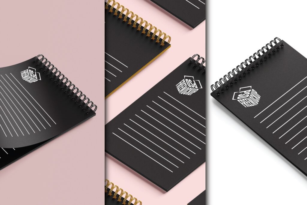 A minimalist spiral notebook mockup PSD displays a black spiral notebook with a golden spiral binder and a white rectangular PSD FREEMOCKUP box logo on the cover. White ruled lines are also visible. The notebook is presented on a soft pink background.