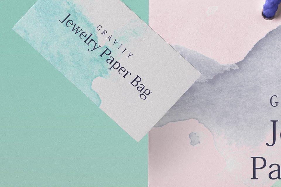A "PSD Shopping Bag Mockup" featuring a white tag with "Gravity Jewelry Paper Bag" text and a teal watercolor splash, overlapping a pink bag with a similar watercolor design, against a teal and pink background.