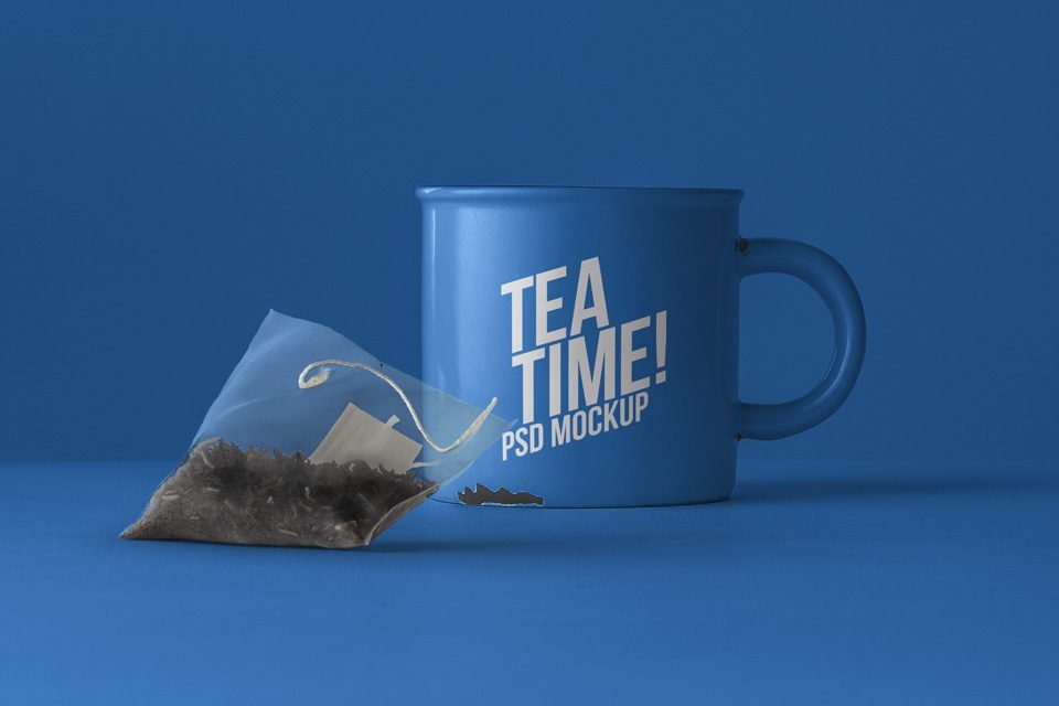 A blue tea mug with white "TEA TIME! PSD MOCKUP" text sits on a blue surface next to a clear, pyramid-shaped tea bag filled with dark tea leaves.